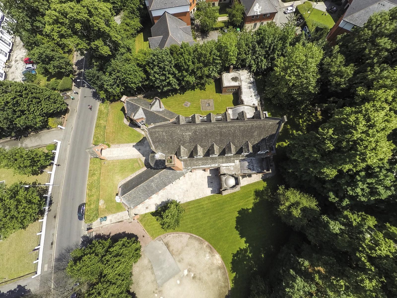 Overhead aerial view of the building's slate roofs and dormers among trees and lawns, with the road running alongside