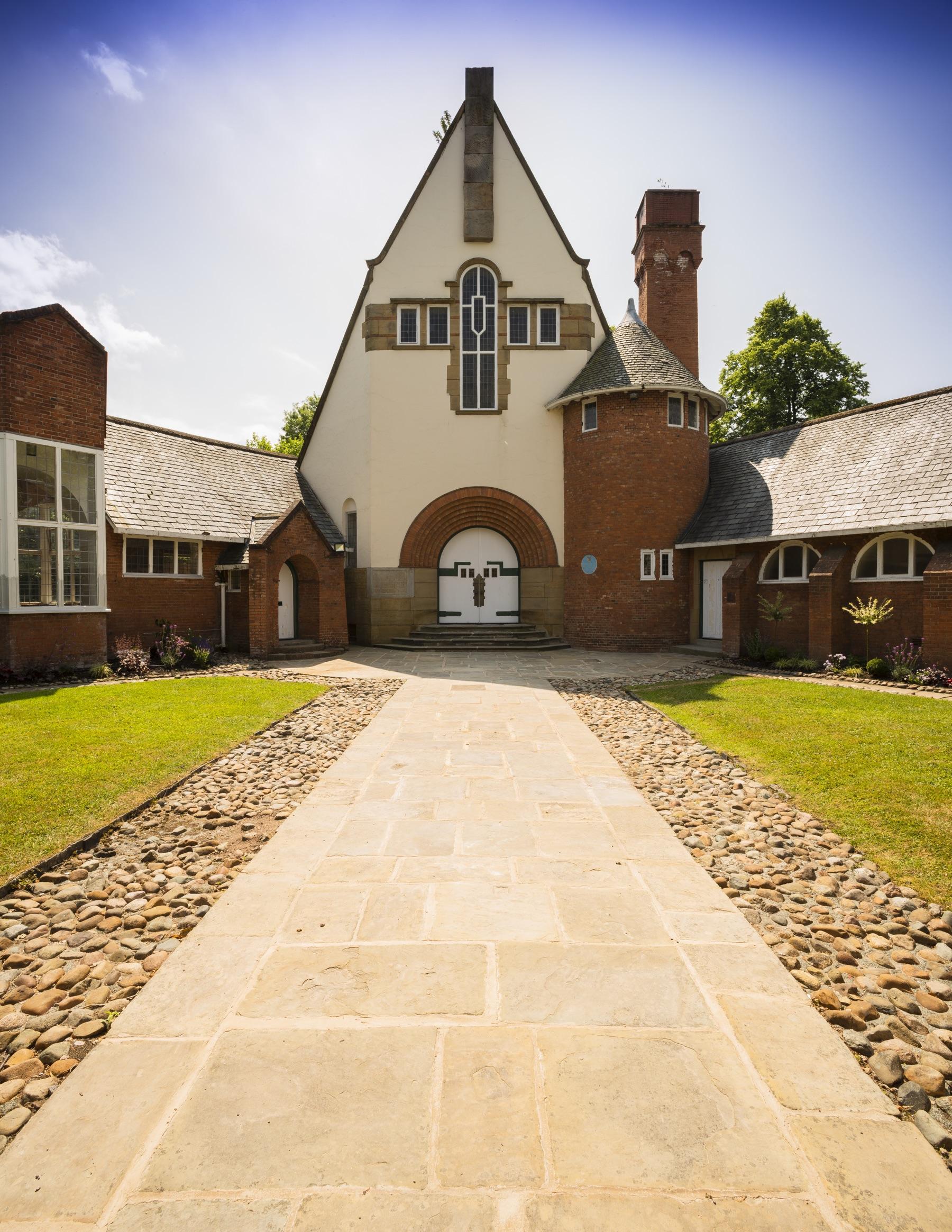 The front of the building: a white rendered gable with a cross-shaped stone window above an arched brick doorway, reached by a flagstone path between lawns